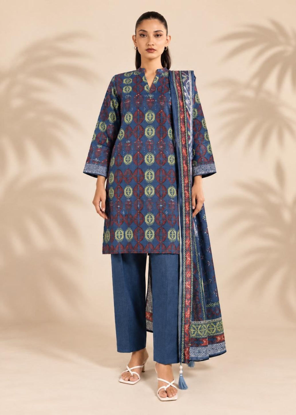 A woman modeling a three-piece traditional outfit consisting of a blue patterned kurta, matching blue trousers, and a coordinating dupatta, set against a beige background with palm leaf shadows.