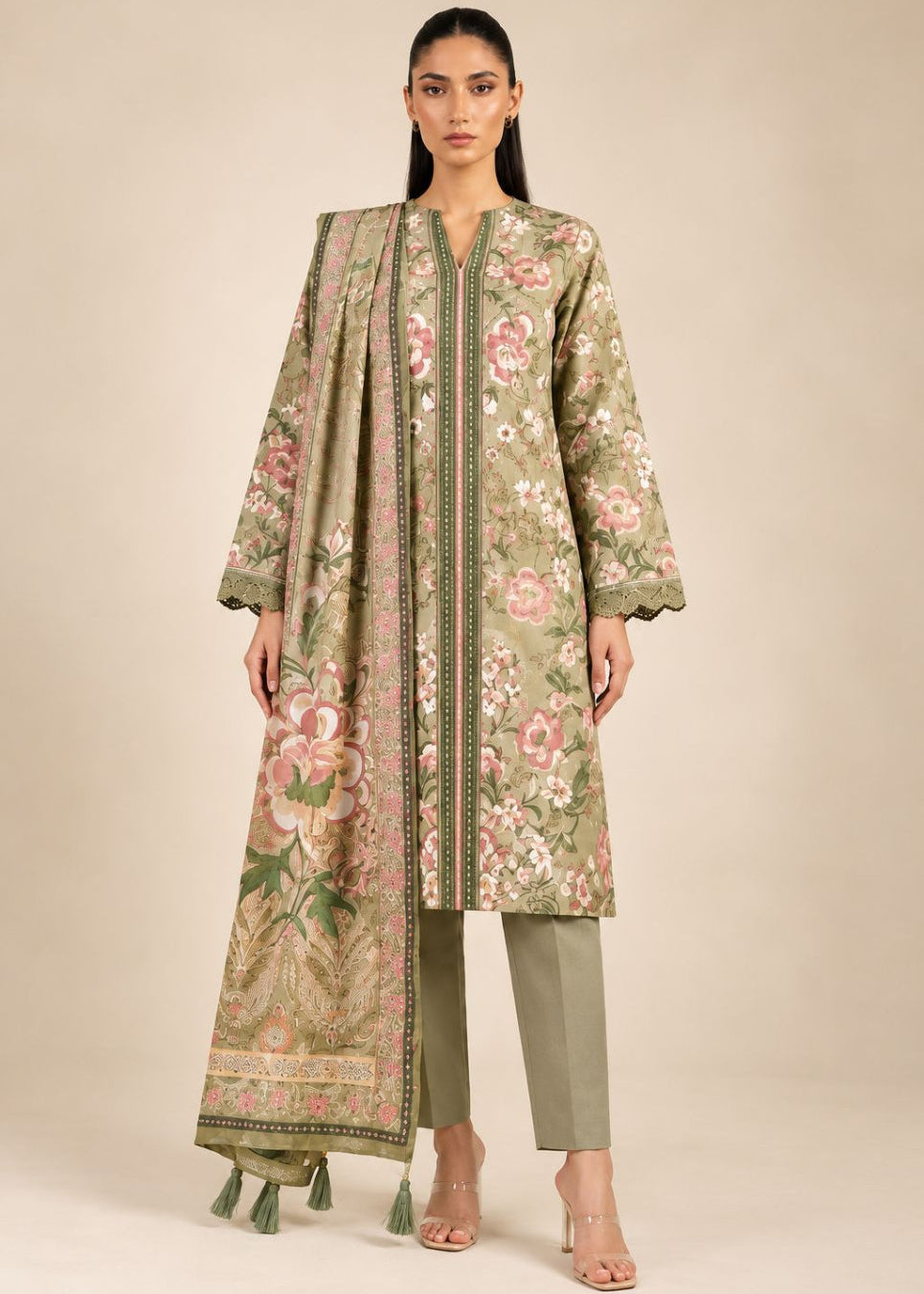 A woman modeling a traditional three-piece outfit featuring a long-sleeved printed tunic in sage green with pink and beige floral motifs, paired with matching sage green trousers and a coordinating dupatta with tassels, set against a solid beige studio background.