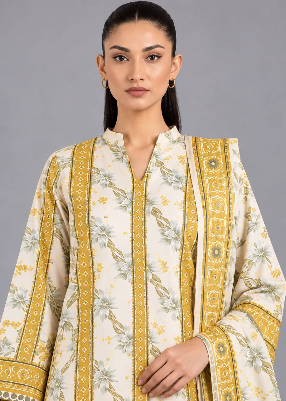 A woman modeling a cream-colored traditional kurta with yellow-gold vertical borders and green floral patterns, set against a solid gray studio background.