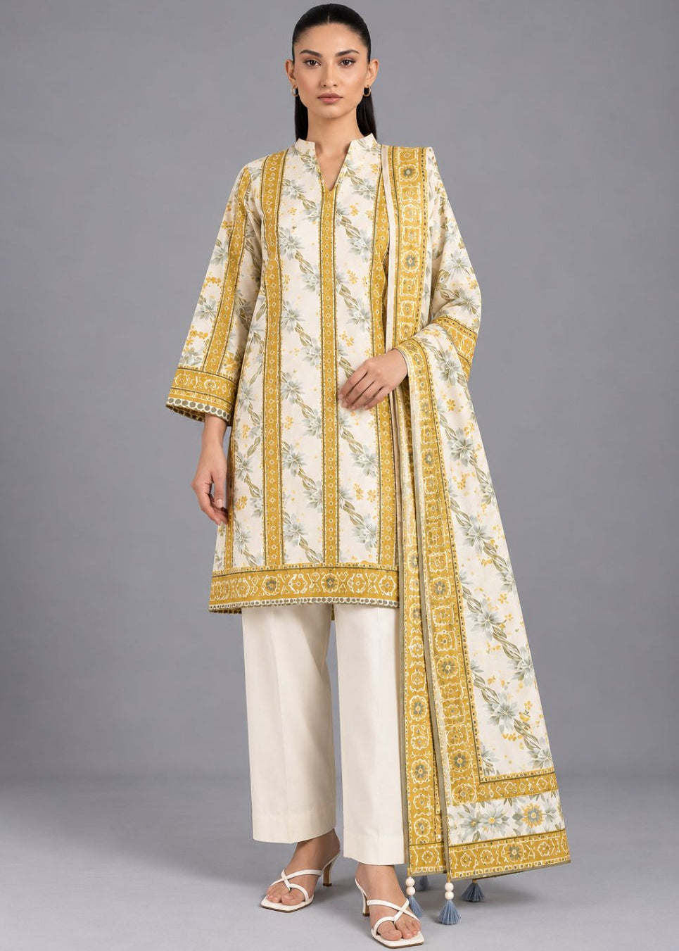 A woman modeling a traditional three-piece salwar kameez suit set. The set features a cream-colored kurta with intricate gold and green floral patterns and gold borders, paired with matching cream wide-leg trousers and a coordinating dupatta.
