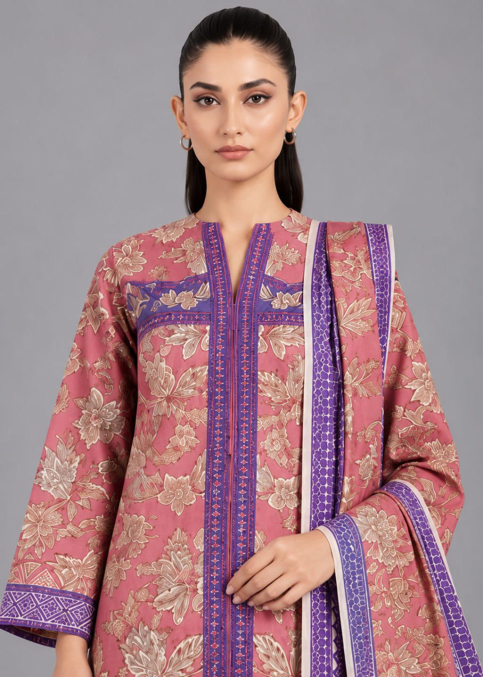 A woman modeling a pink floral traditional outfit with purple border details and a matching dupatta against a solid gray studio background.