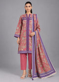 A woman modeling a traditional three-piece salwar kameez suit in dusty pink with purple borders and beige floral patterns, paired with matching pink pants and a coordinating dupatta, against a solid gray studio background.