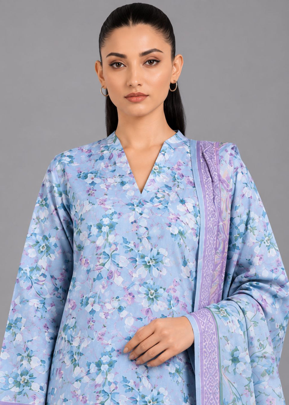 A woman modeling a light blue floral kurta with a matching purple patterned dupatta against a solid gray studio background.