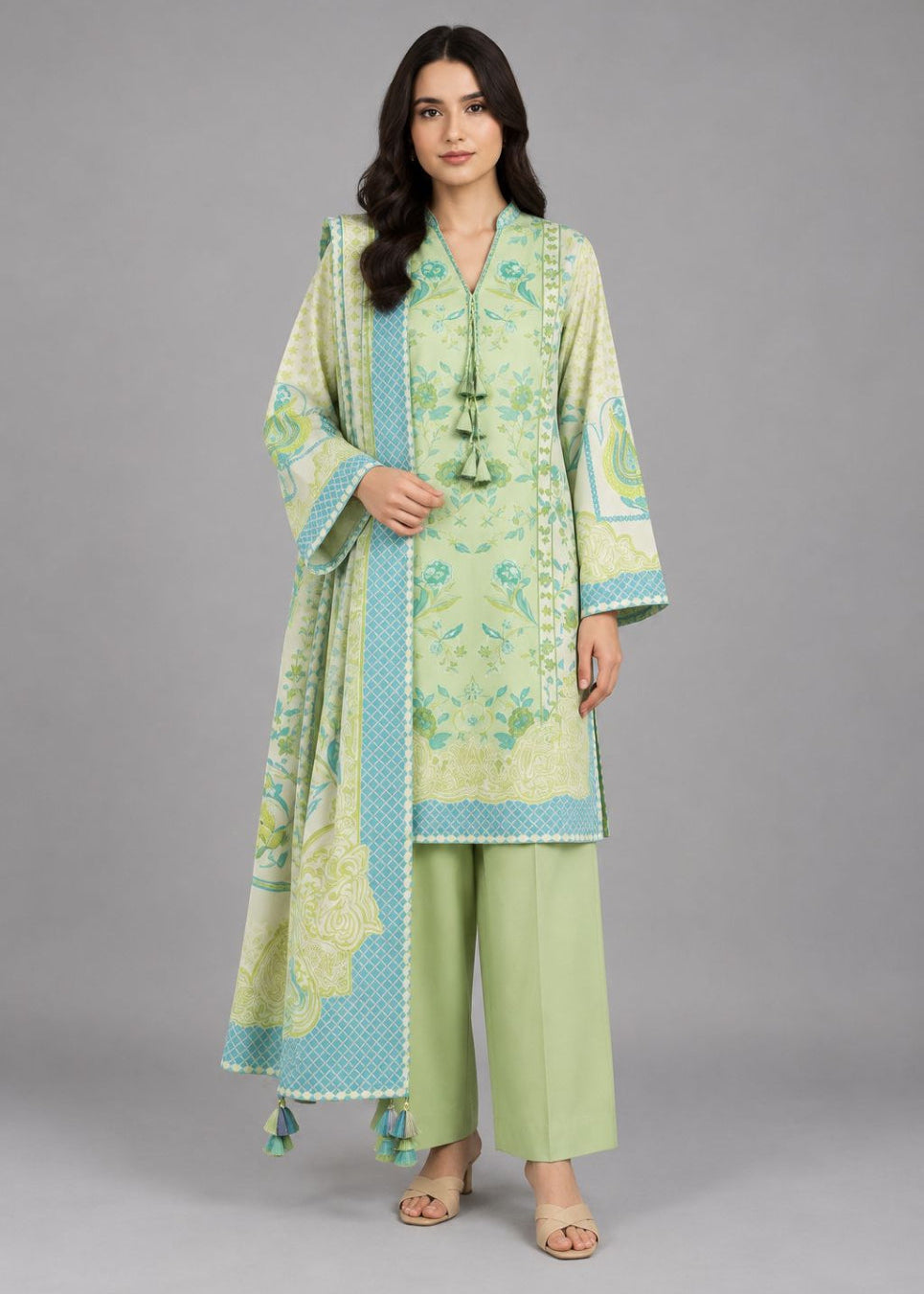 A woman modeling a light green and turquoise printed traditional three-piece outfit with a matching dupatta, paired with light green pants, against a solid gray studio background.