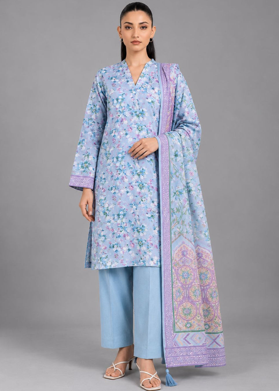A woman modeling a light blue floral printed kurta set with matching light blue pants and a lavender-purple patterned dupatta, photographed against a solid gray studio background.