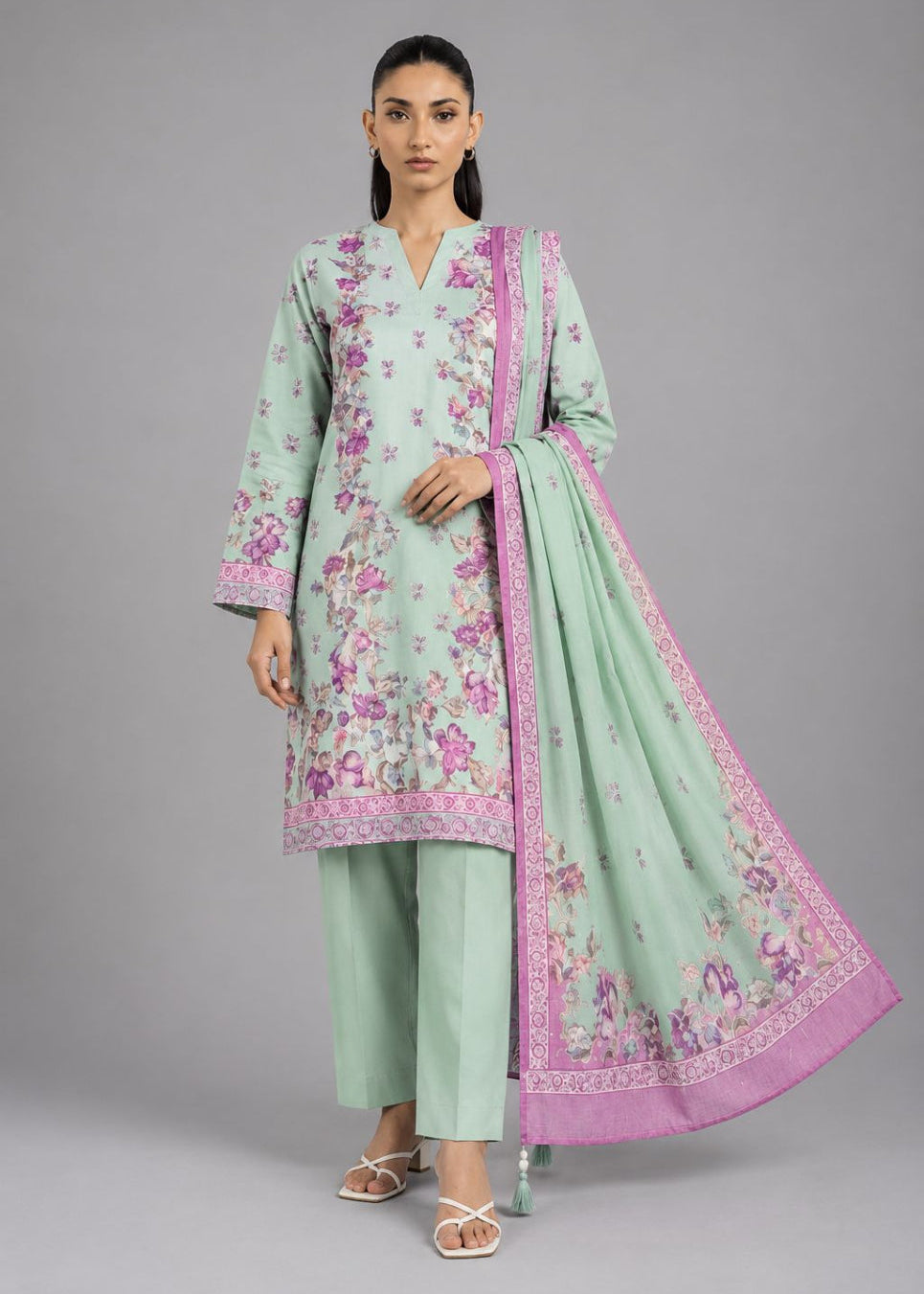 A woman modeling a mint green three-piece traditional outfit with pink floral embroidery and a matching pink-bordered dupatta, photographed against a plain gray studio background.