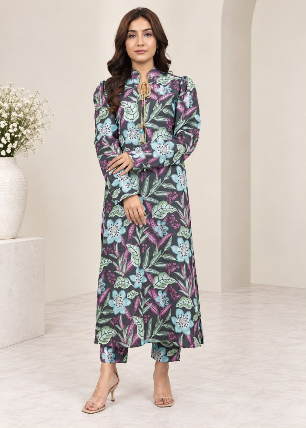 A woman models a long-sleeved, high-neck maxi dress with a colorful floral and leaf pattern in purple, green, and light blue, styled with matching cropped pants and beige high-heeled sandals.