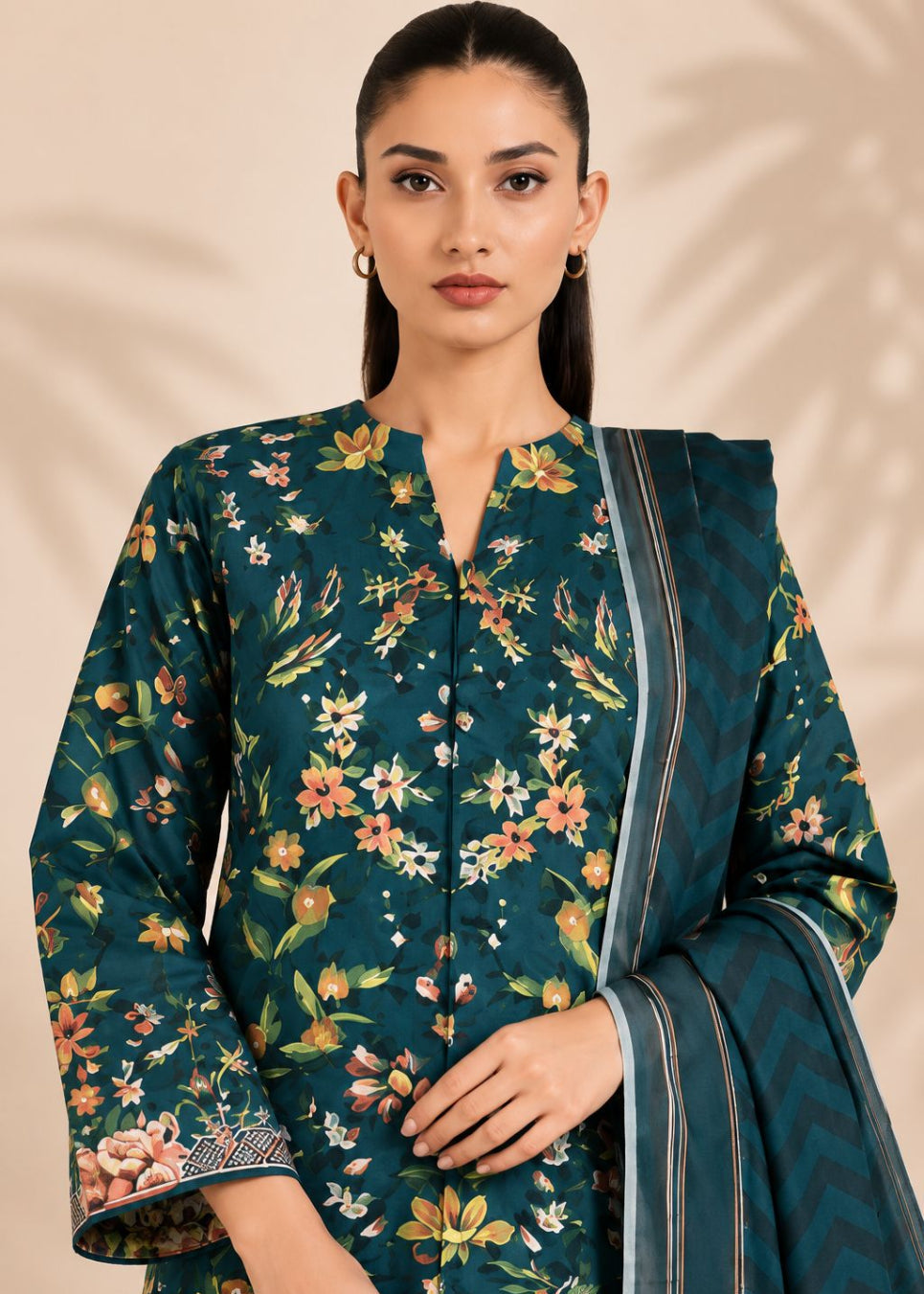 A woman modeling a dark teal floral salwar kameez suit with a matching patterned dupatta, set against a warm beige background with soft leaf shadows.