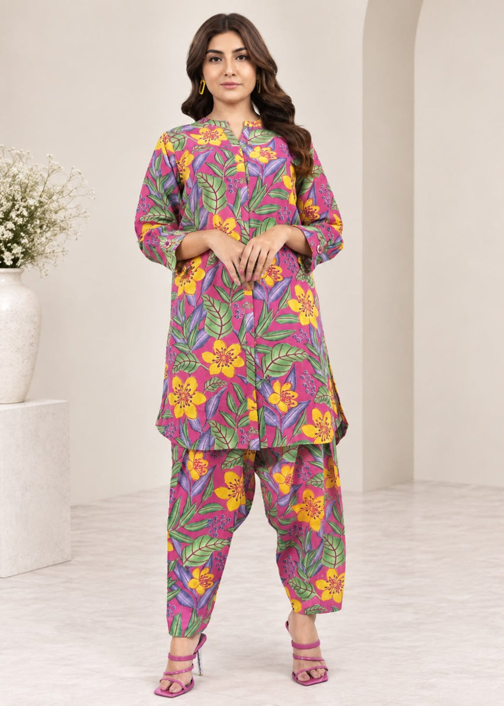 A woman modeling a colorful printed kurta and pant set in a bright, modern studio setting. The outfit features a vibrant floral and leaf pattern in pink, purple, yellow, and green.