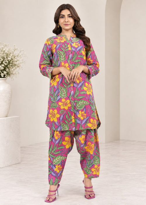 A woman modeling a colorful printed kurta and pant set in a bright, modern studio setting. The outfit features a vibrant floral and leaf pattern in pink, purple, yellow, and green.