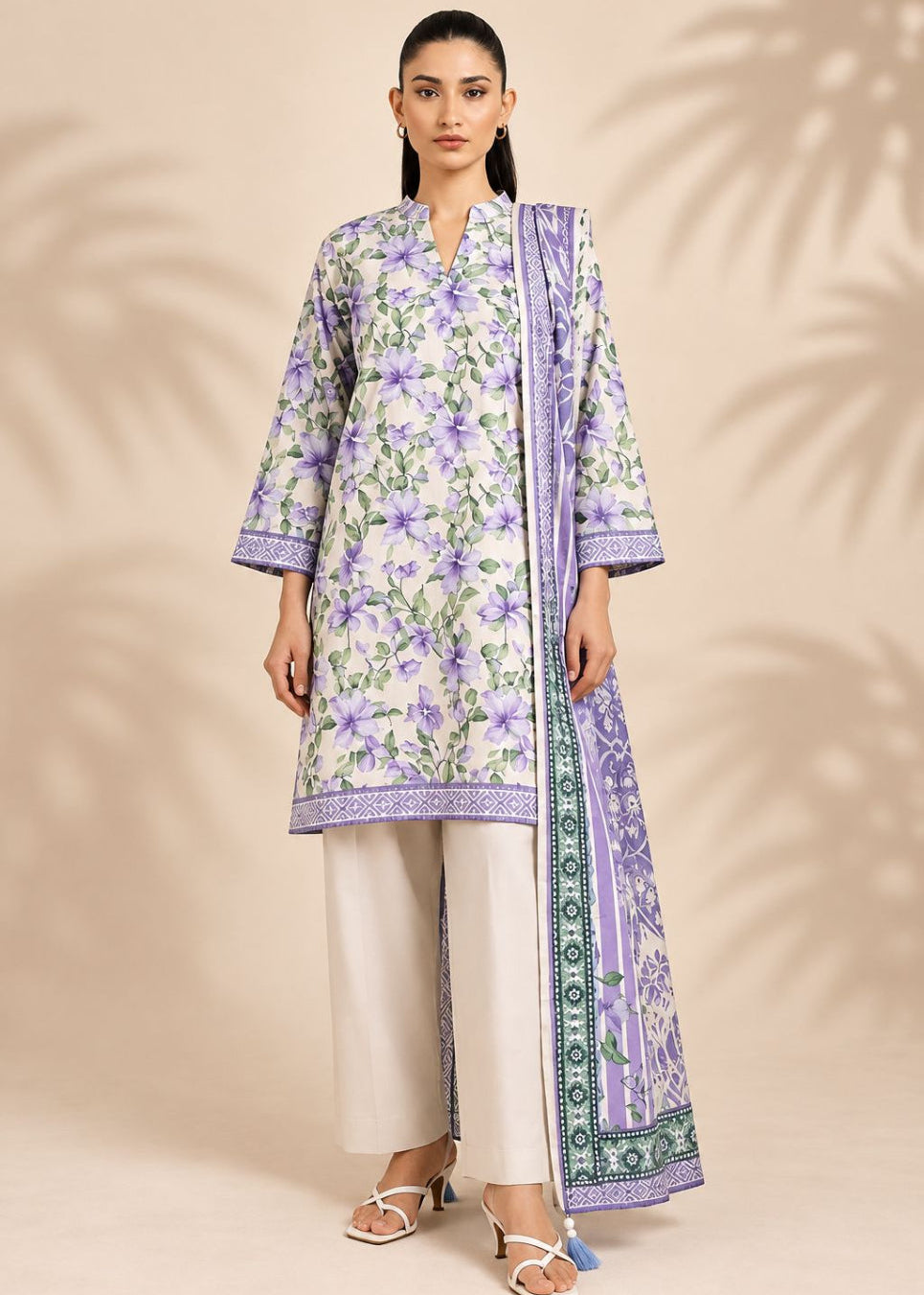 A woman models a traditional three-piece salwar kameez suit set. The set features a lavender and green floral print kurta, cream-colored wide-leg trousers, and a matching printed dupatta with a green border, set against a beige background with leaf shadows.