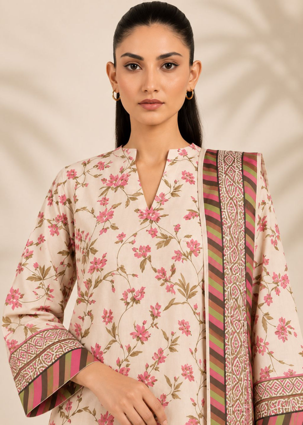 A woman modeling a cream-colored floral kurta with pink and green floral patterns, featuring wide sleeves with striped and geometric borders, paired with a matching patterned dupatta.