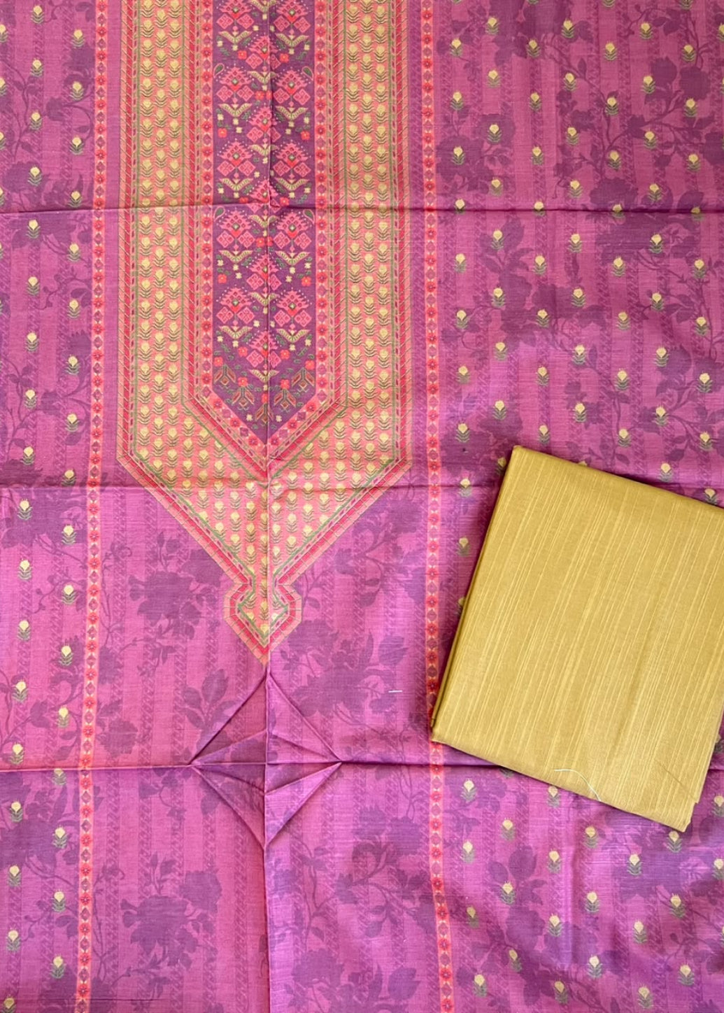 A pink and purple patterned fabric laid flat, featuring a central embroidered panel and small gold dot motifs, with a folded matching yellow-gold fabric piece placed in the lower right corner.