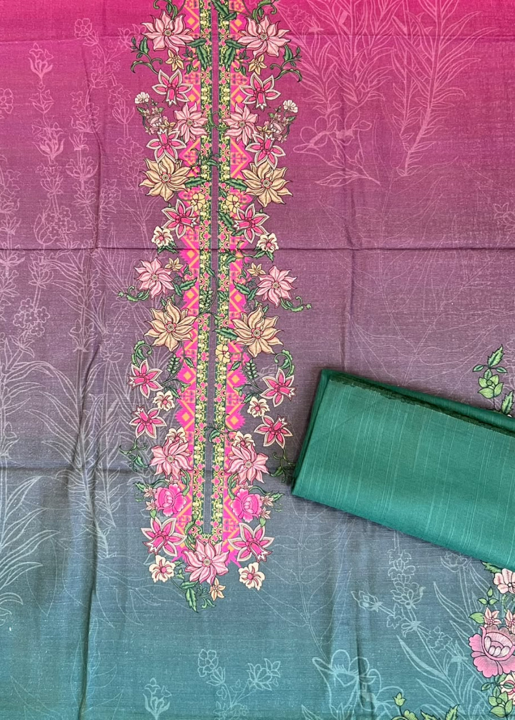 A folded and flat-laid silk saree fabric with a gradient design transitioning from magenta pink to teal green, decorated with a central floral embroidery panel and pink floral motifs.