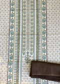 A folded dark brown fabric piece, likely a dupatta or scarf, placed on top of a larger patterned textile. The larger textile has a cream background with a dense small floral print and two vertical decorative borders in teal, green, and gold.