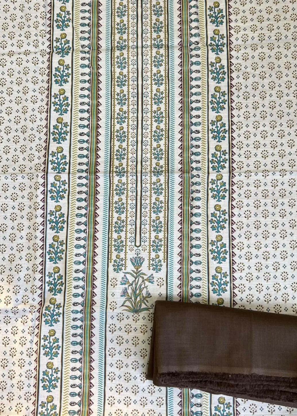 A folded dark brown fabric piece, likely a dupatta or scarf, placed on top of a larger patterned textile. The larger textile has a cream background with a dense small floral print and two vertical decorative borders in teal, green, and gold.