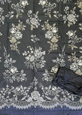 A black fabric garment or textile laid flat on green artificial turf, featuring an allover white and beige floral print with a decorative lace-like border. A folded black matching piece and a person's feet in sandals are visible at the bottom, along with a yellow shopping bag in the lower right.