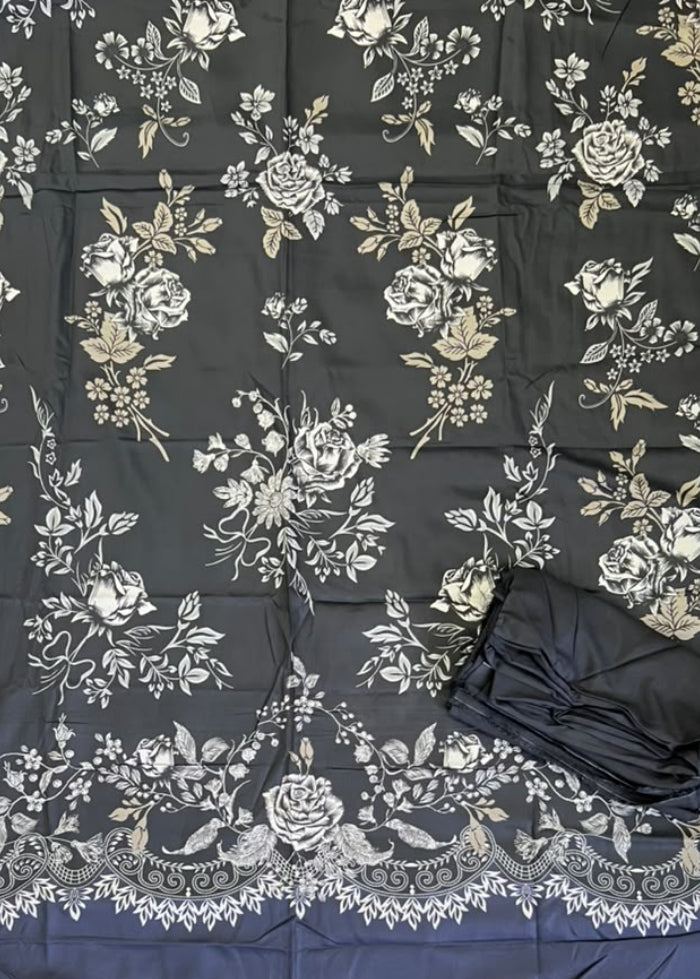 A black fabric garment or textile laid flat on green artificial turf, featuring an allover white and beige floral print with a decorative lace-like border. A folded black matching piece and a person's feet in sandals are visible at the bottom, along with a yellow shopping bag in the lower right.