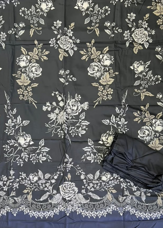 A black fabric garment or textile laid flat on green artificial turf, featuring an allover white and beige floral print with a decorative lace-like border. A folded black matching piece and a person's feet in sandals are visible at the bottom, along with a yellow shopping bag in the lower right.