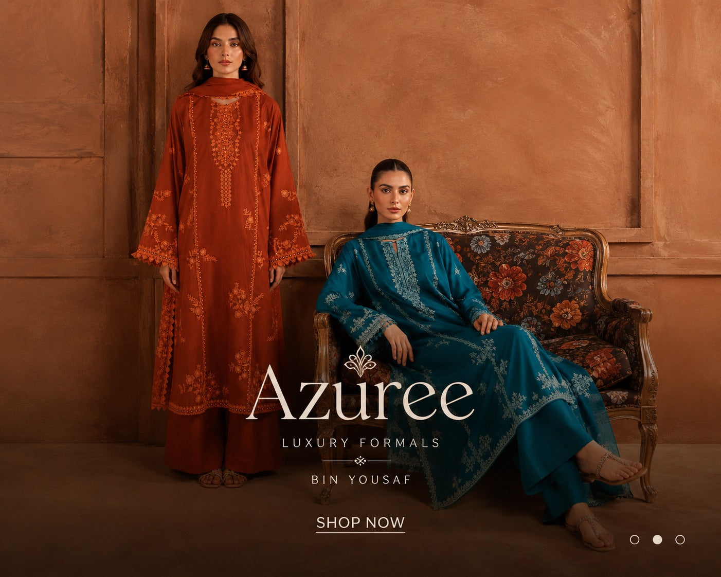 Two models showcase luxury formal ethnic wear from Azuree. One stands in a rust-orange kurta with intricate embroidery, and the other sits on an ornate floral sofa in a teal-blue kurta with matching dupatta.