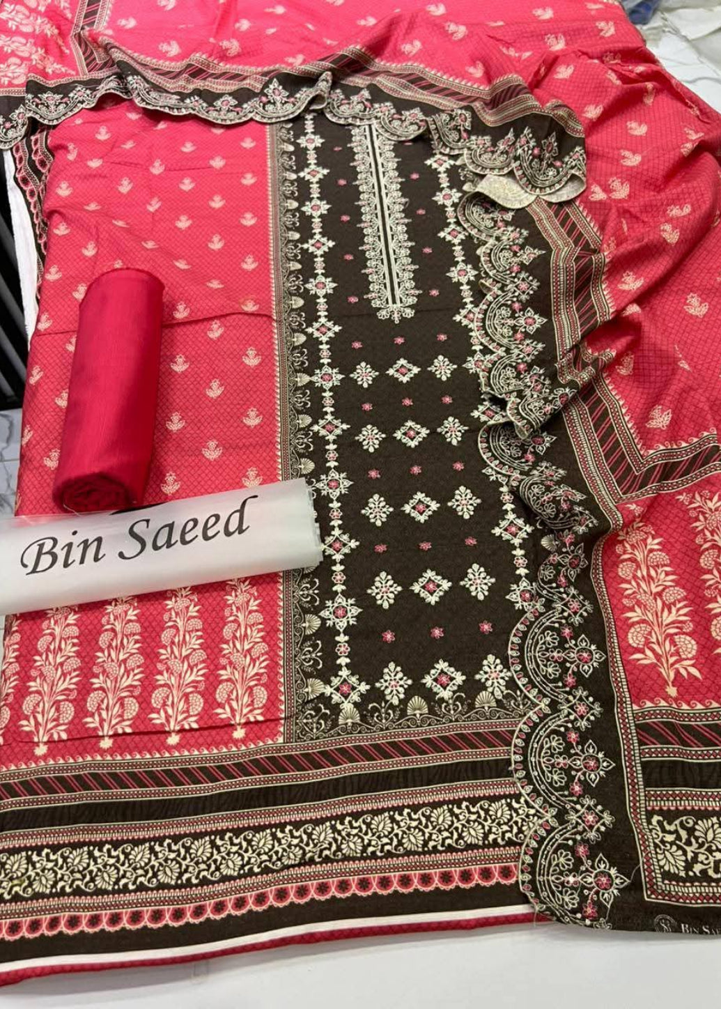 A folded and rolled traditional South Asian salwar suit material set by Bin Saeed, featuring a vibrant pink fabric with gold floral prints, a dark brown base with intricate gold and green geometric embroidery, and a matching pink dupatta.