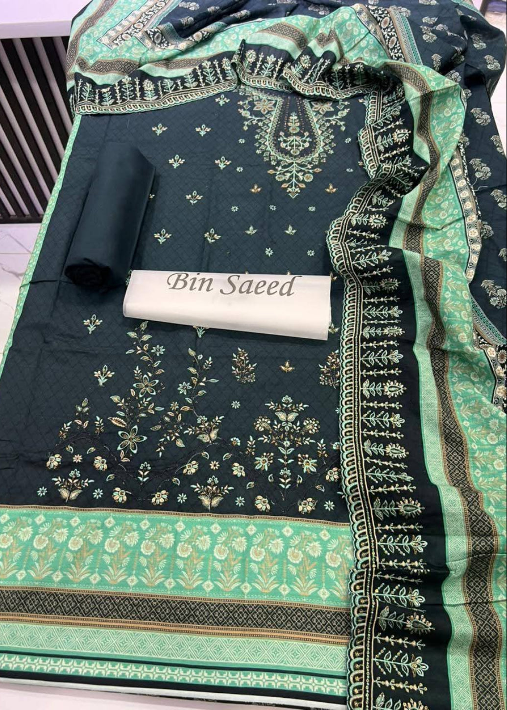 A dark green and mint green embroidered fabric set with a matching dupatta, featuring intricate floral and paisley patterns, a rolled fabric piece, and a white label reading 'Bin Saeed'.