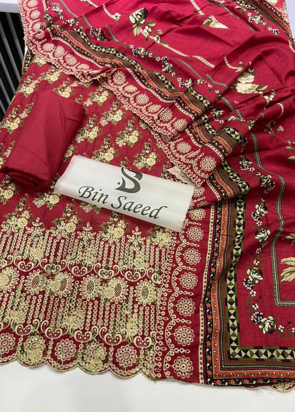A red traditional South Asian fabric set with intricate gold and multicolor floral embroidery, a matching red fabric piece folded on top, and a white label printed with the brand name 'Bin Saeed'.