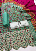 A folded and rolled green floral salwar suit fabric set with a matching pink dupatta and ornate embroidered border, displayed on a white surface with a Bin Saeed label.