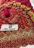 A deep maroon red ethnic suit fabric set with intricate gold, green, and orange floral embroidery and mirror work, featuring a matching dupatta, rolled fabric, and a white brand label reading 'Bin Saeed'.