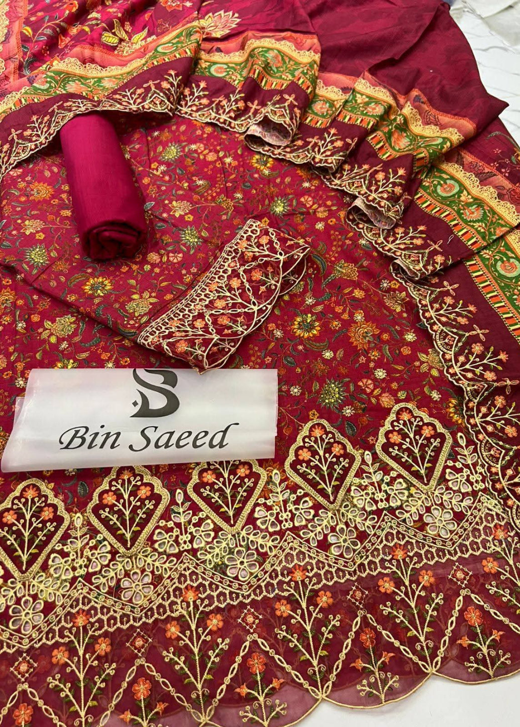 A deep maroon red ethnic suit fabric set with intricate gold, green, and orange floral embroidery and mirror work, featuring a matching dupatta, rolled fabric, and a white brand label reading 'Bin Saeed'.