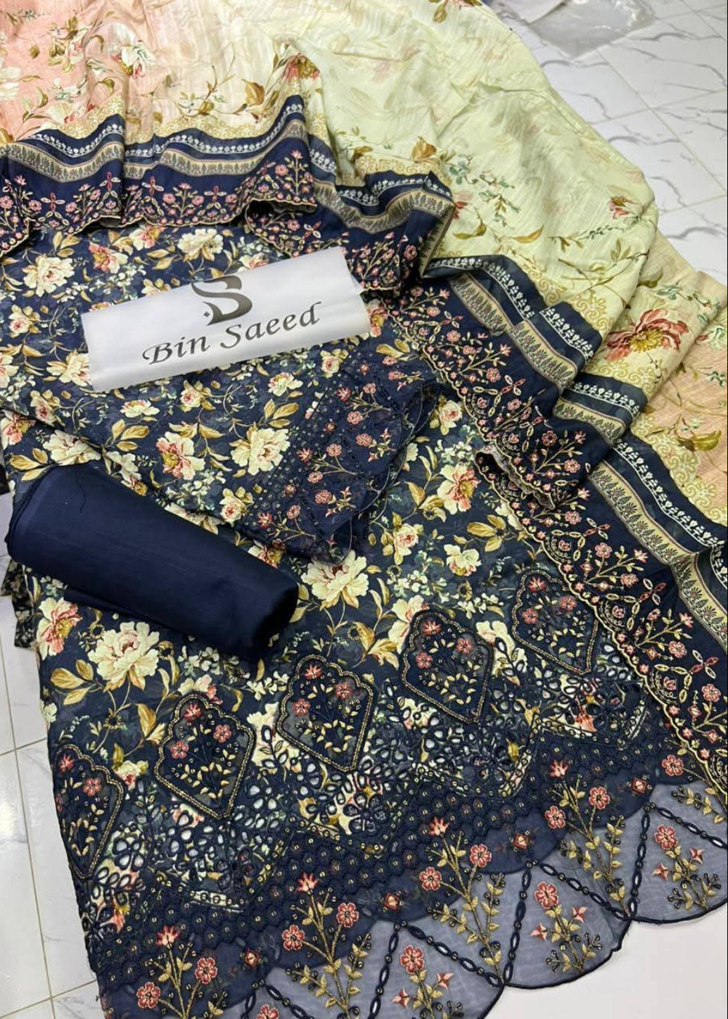 A folded and draped navy blue floral salwar suit fabric set with matching printed dupatta and a rolled fabric piece, displayed on a light tiled floor. A white label reading 'Bin Saeed' is placed on top.