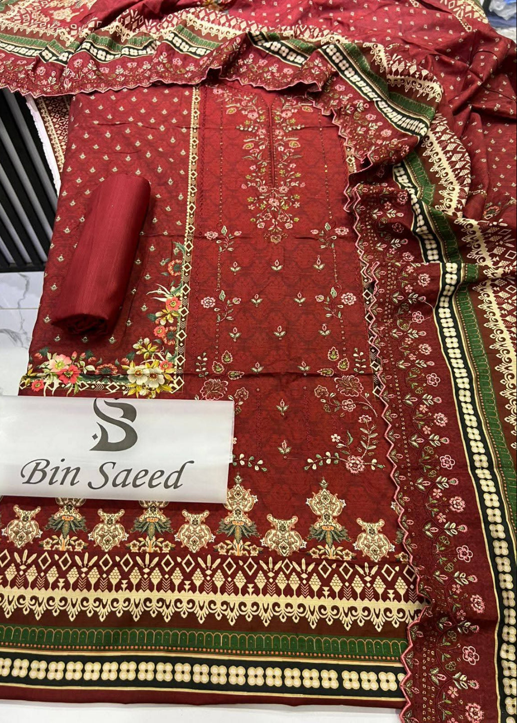 A folded and draped red ethnic fabric set with intricate gold and green floral embroidery and border patterns, featuring a matching rolled fabric piece and a white label reading 'Bin Saeed'.