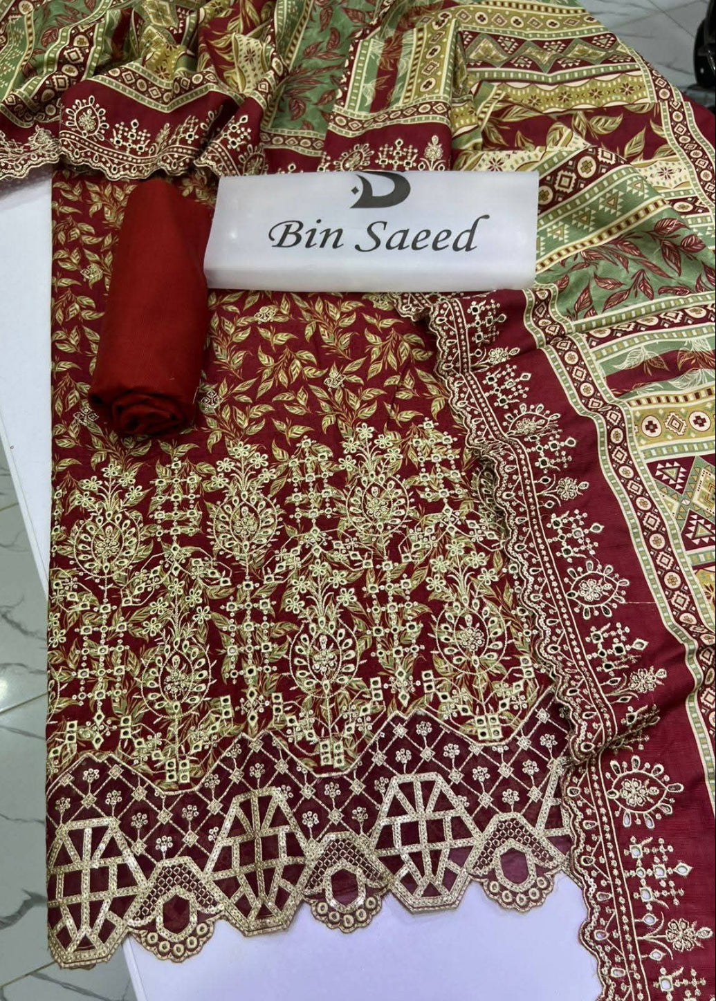 A traditional embroidered fabric set in deep red, featuring intricate gold and beige floral patterns, a matching rolled fabric piece, and a white label reading 'Bin Saeed'.