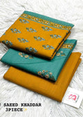 Three folded pieces of Bin Saeed Khaddar fabric in mustard yellow and teal green with floral motifs, arranged on a white faux fur background with a brand tag and text overlay.