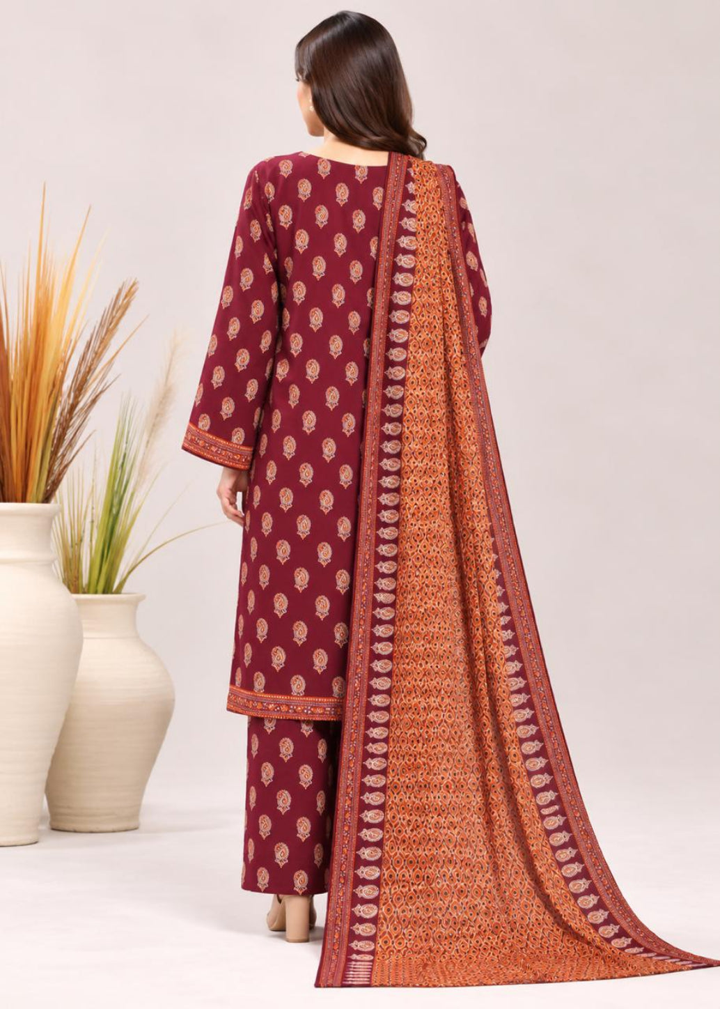 A woman shown from the back wearing a three-piece salwar kameez suit in maroon and orange. The kurta features small beige motifs, the pants are maroon, and a matching orange dupatta with a detailed border is draped over her shoulder.