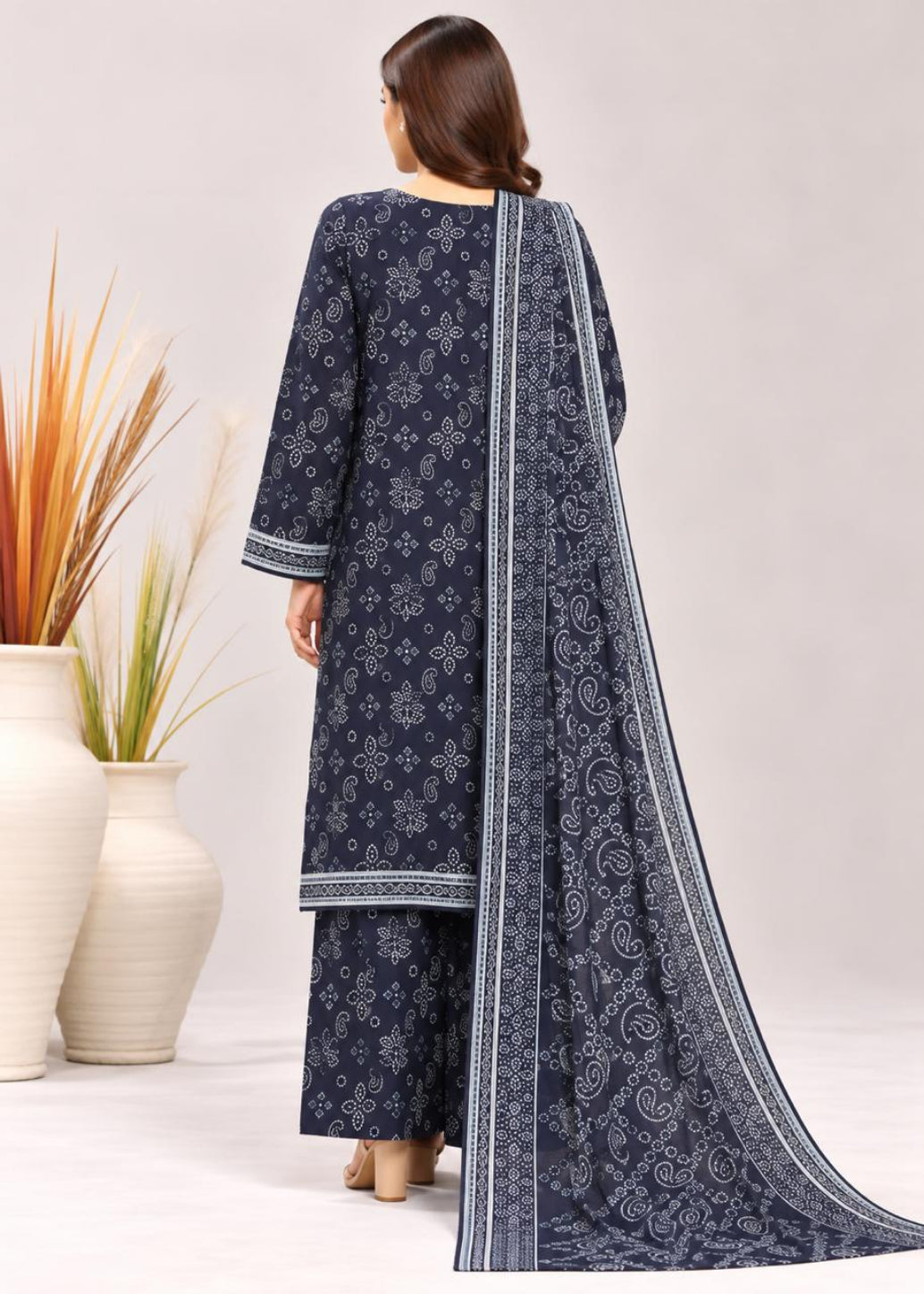 A woman shown from the back wearing a navy blue three-piece salwar kameez suit with white printed patterns and a matching dupatta. The set is photographed against a light gray studio background with decorative white vases containing dried grass on the left.