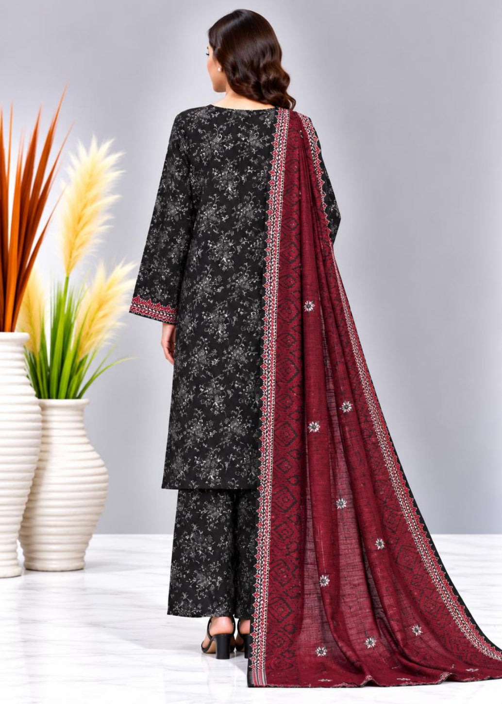 A woman shown from the back wearing a black floral printed kurta set with a deep red dupatta featuring a white border and small floral motifs, photographed in a studio with decorative vases on the left.