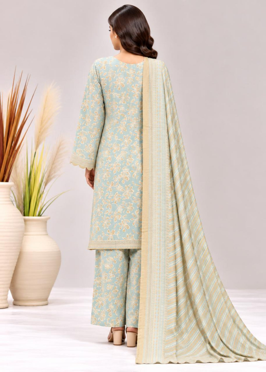 A woman shown from the back wearing a light blue and beige floral salwar kameez suit with a matching striped dupatta, standing in a studio with decorative vases.