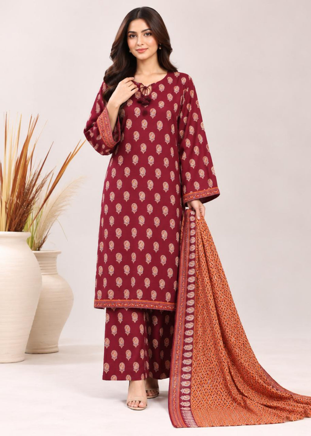 A woman models a three-piece salwar kameez suit in maroon with beige block prints, paired with a rust-orange printed dupatta, set against a neutral studio background with decorative vases.