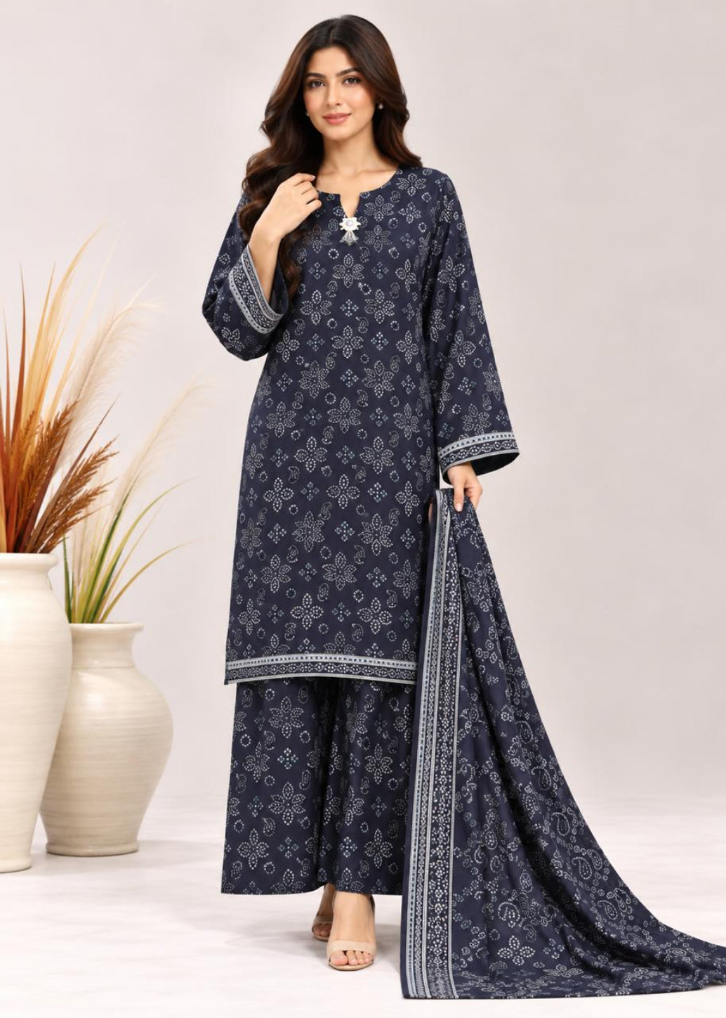 A woman models a navy blue three-piece salwar kameez suit with white geometric prints and border details, including a matching dupatta, set against a light beige studio background with decorative vases.