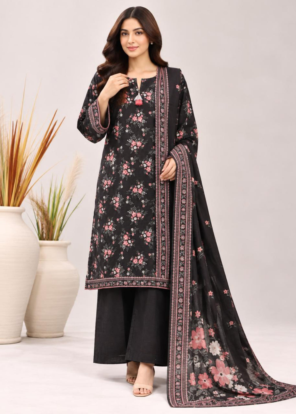 A woman models a black three-piece salwar kameez suit with pink floral prints and a matching dupatta, styled with black wide-leg pants. The set features intricate border detailing and is photographed against a neutral studio backdrop with decorative vases.