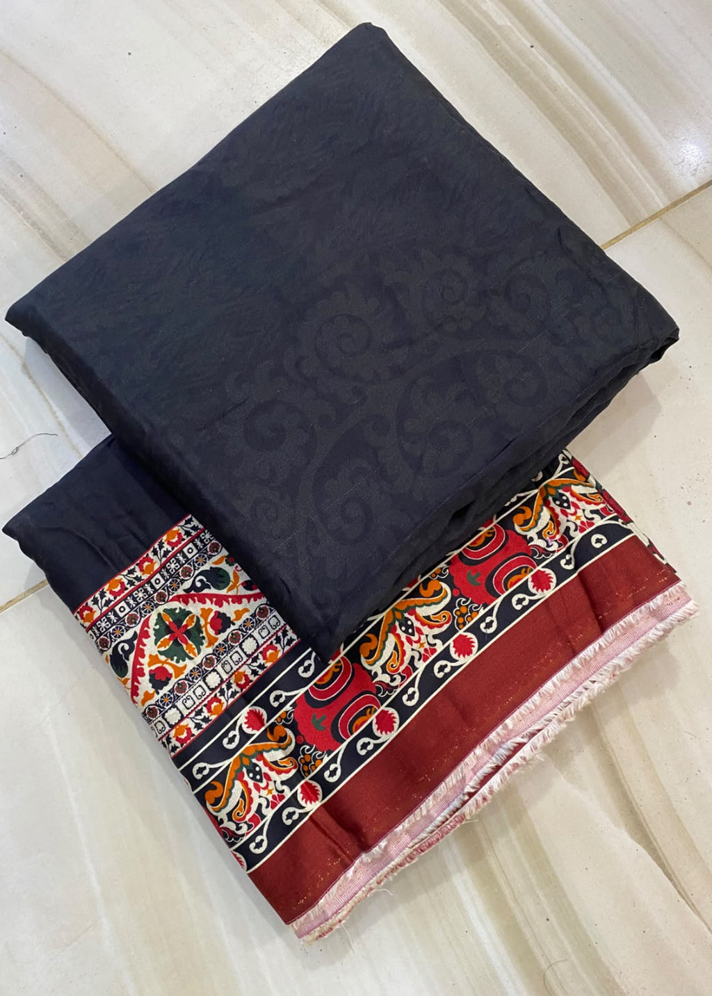 A folded dark charcoal gray fabric piece with a subtle jacquard pattern, layered over a matching dark fabric and a red, black, and multicolor patterned border fabric, displayed on a light beige marble floor.