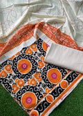 A folded and draped saree fabric set on a green grass-like surface, featuring a white base with pink floral embroidery, a wide orange border, and a black central panel with orange, pink, and white circular motifs.
