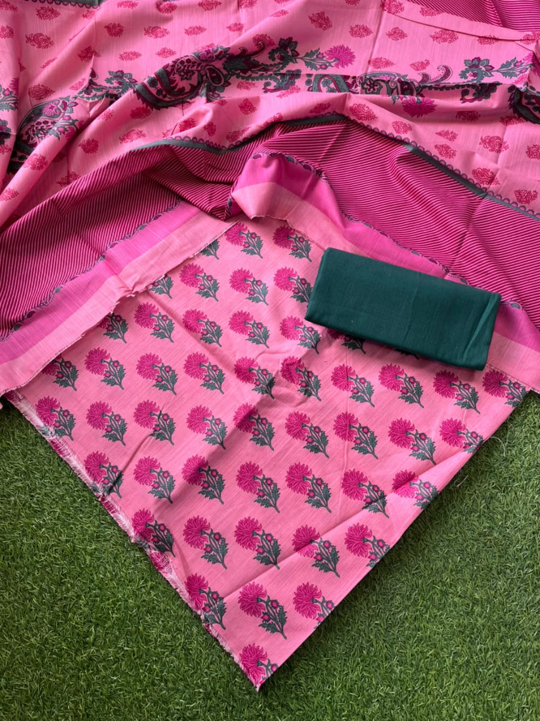 Pink floral printed fabric with a matching striped border and a folded dark green fabric piece placed on top, laid out on green grass.