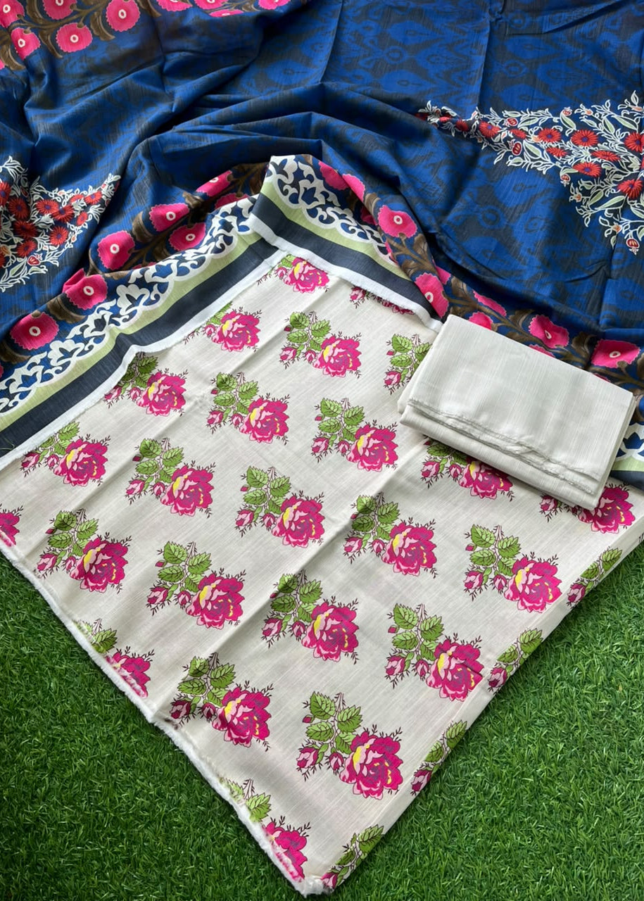 A folded floral fabric set laid out on green artificial grass, featuring a cream-colored base with pink and green floral print, a matching blue patterned dupatta with red and white floral borders, and a folded light gray fabric piece.