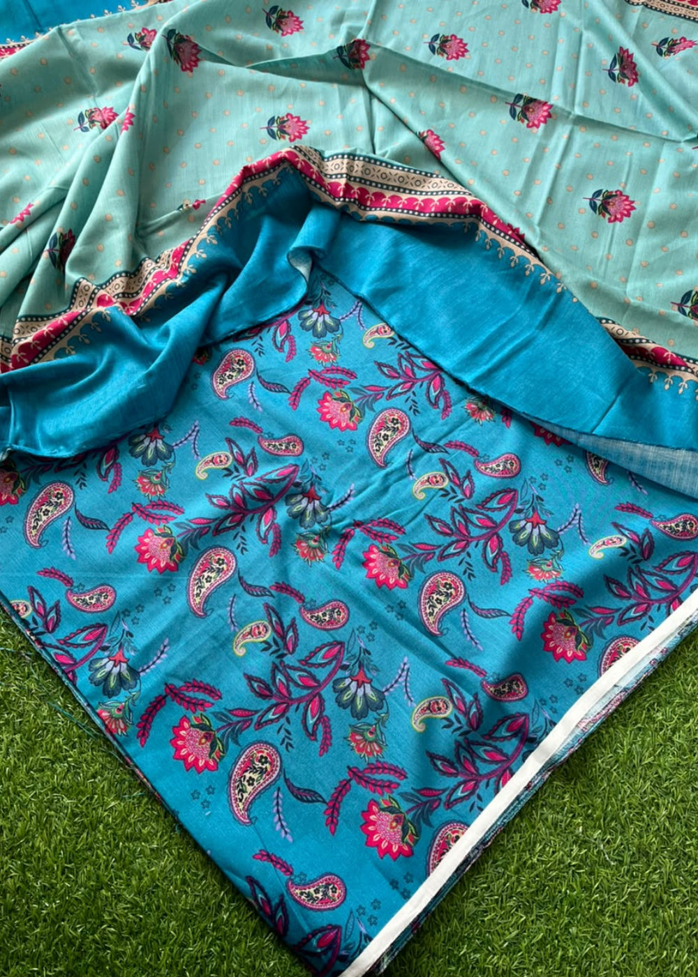 A folded and draped fabric set, likely a sari or scarf, featuring a turquoise blue base with intricate paisley and floral block prints in pink, red, and green, paired with a light mint green border section.