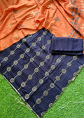 A navy blue fabric garment laid flat on green artificial grass, with an orange patterned dupatta draped across the top. The navy fabric has repeated circular motifs and a fringed edge, and a folded matching fabric piece sits on top.