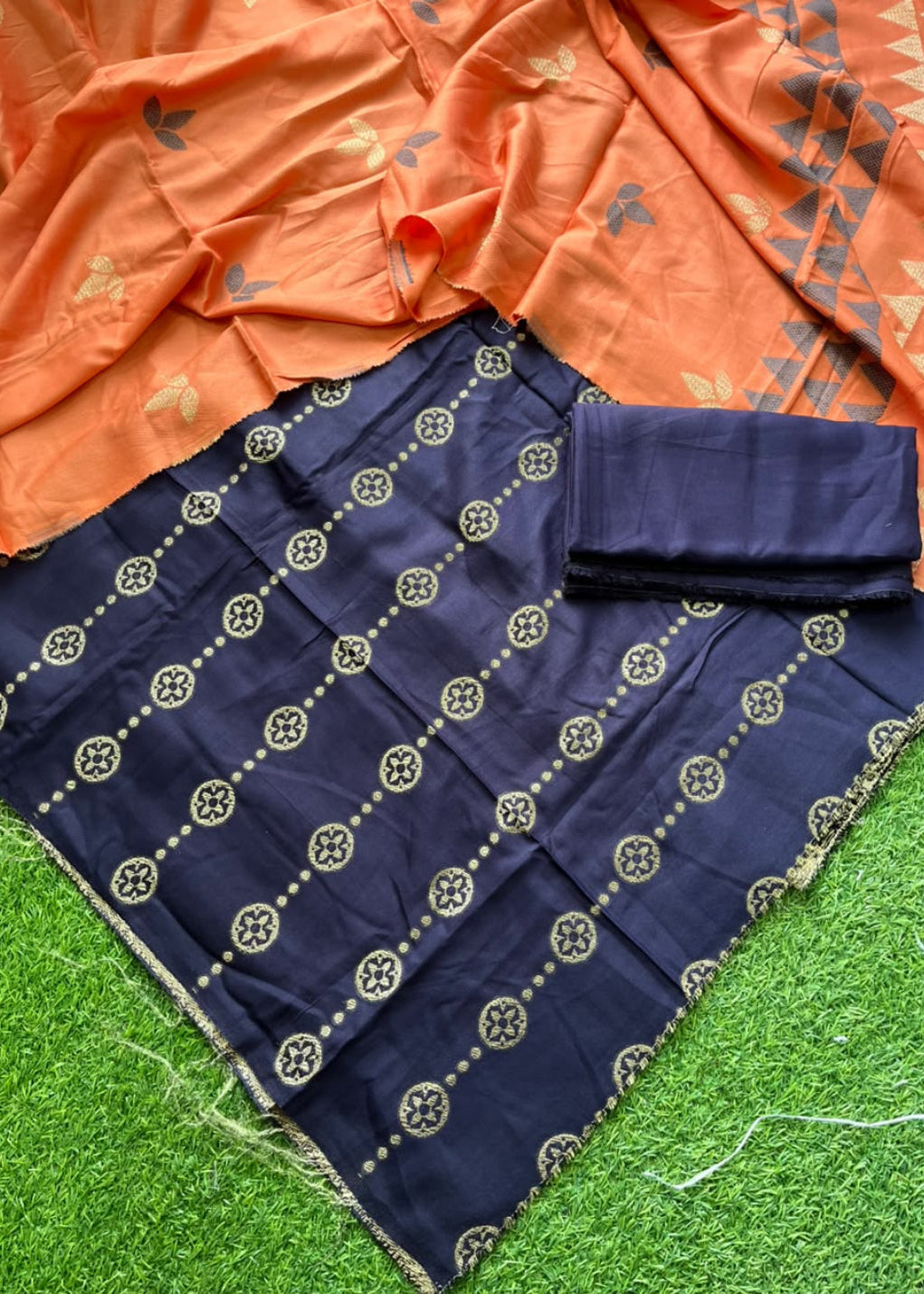 A navy blue fabric garment laid flat on green artificial grass, with an orange patterned dupatta draped across the top. The navy fabric has repeated circular motifs and a fringed edge, and a folded matching fabric piece sits on top.