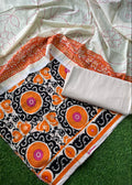 A folded and draped saree fabric set on a green grass-like surface, featuring a white base with pink floral embroidery, a wide orange border, and a black central panel with orange, pink, and white circular motifs.