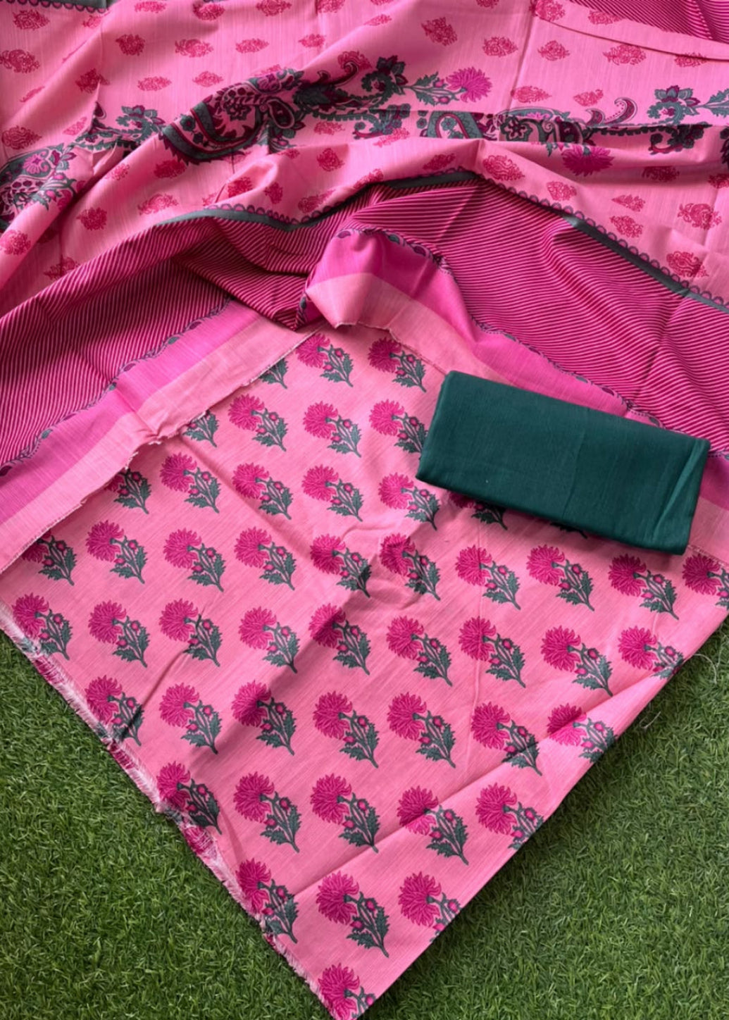Pink floral printed fabric with a matching striped border and a folded dark green fabric piece placed on top, laid out on green grass.